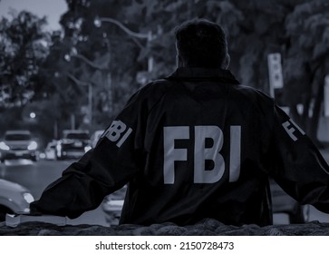 Agente del FBI, un hombre, con un abrigo azul oscuro y el logo del FBI mirando por la calle con coches en el atardecer visto desde atrás.