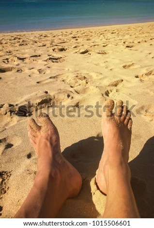 Male feet covered in sand, relaxing on the beach with clear aqua water in the background