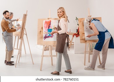 Male And Female Adult Students In Aprons Painting Together On Easels In Art Class