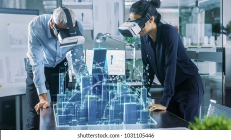 Male And Female Architects Wearing  Augmented Reality Headsets Work With 3D City Model. High Tech Office Professional People Use Virtual Reality Modeling Software Application.