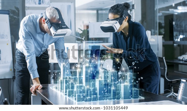 Male and Female
Architects Wearing  Augmented Reality Headsets Work with 3D City
Model. High Tech Office Professional People Use Virtual Reality
Modeling Software
Application.