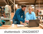 Male And Female Carpenters Working In Woodwork Workshop Using Laptop Together