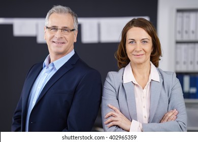 Male And Female Confident Middle Aged Business People Grinning While Standing In Front Of Wall Of Charts And Bookshelf In Office