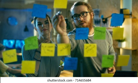 Male and Female Designers Discuss and Outline They're Project with Sticky Notes on a Glass Wall. They're Young and Creative People Who Work in a Stylish Loft Space.