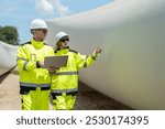 Male and female engineer in uniform with helmet safety using laptop inspection and maintenance large wind turbine blade in wind farms to generate electrical energy, Renewable energy.