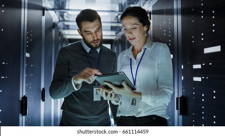 Male And Female IT Engineers In Data Center / Server Room, They Are Talking. One Of Them Holds Tablet Computer.