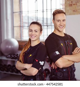 Male and female fitness instructors stand together while wearing ems outfits in exercise studio near tall windows