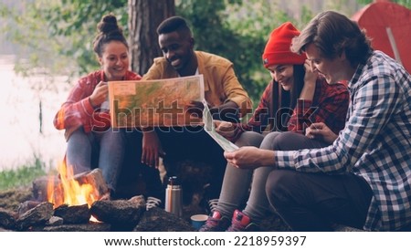 male and female friends tourists are sitting around fire and studying maps during hike in forest in summer. Young people are wearing casual clothing.