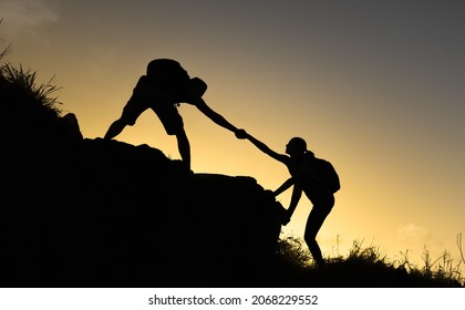 Male And Female Hiker Silhouette Helping Each Other Climb Up A Mountain. Teamwork Concept. 