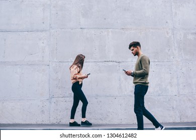 Male And Female Hipsters Walking Outdoors Looking At Their Smartphones Ignore Real Life, Girl And Guy Over Using Concentrated On Using Mobile Phone For Chatting And Networking Addicted To Gadgets