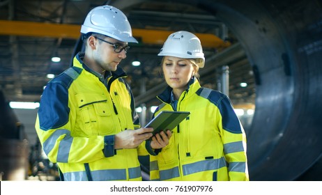 Male and Female Industrial Engineers in Hard Hats Discuss New Project while Using Tablet Computer. They're Making Calculated Engineering Decisions.They Work at the Heavy Industry Manufacturing Factory