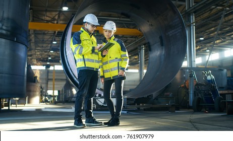 Male and Female Industrial Engineers in Hard Hats Discuss New Project while Using Tablet Computer. They're Making Calculated Engineering Decisions.They Work at the Heavy Industry Manufacturing Factory