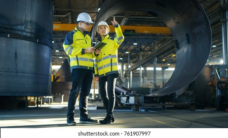 Male and Female Industrial Engineers in Hard Hats Discuss New Project while Using Tablet Computer. They're Making Calculated Engineering Decisions.They Work at the Heavy Industry Factory.