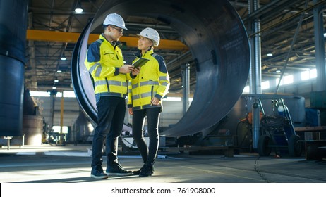 Male and Female Industrial Engineers in Hard Hats Discuss New Project while Using Tablet Computer. They're Making Calculated Engineering Decisions.They Work at the Heavy Industry Manufacturing Factory