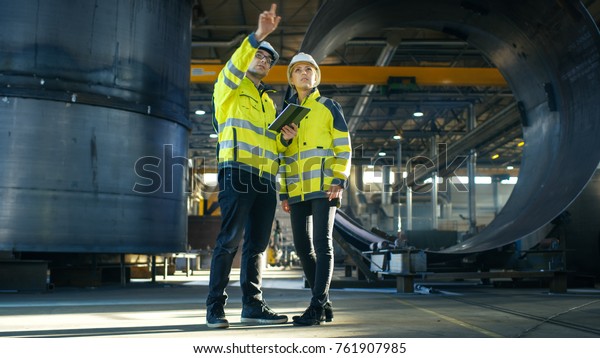 Male and Female Industrial Engineers in Hard Hats\
Discuss New Project while Using Tablet Computer. They\'re Making\
Calculated Engineering Decisions.They Work at the Heavy Industry\
Manufacturing Factory