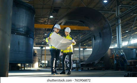 Male And Female Industrial Engineers Look At Project Blueprints While Standing Surround By Pipeline Parts In The Middle Of Enormous Heavy Industry Manufacturing Factory