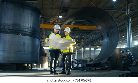 Ingenieure von männlichen und weiblichen Industriebetrieben betrachten Projektkonzepte, während sie in der Mitte der Fabrik für die Herstellung von energiegeladenen Schwerindustrien durch Rohrleitungen umrahmen