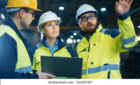 Male And Female Industrial Engineers Talk With Factory Worker While Using Laptop. They Work At The Heavy Industry Manufacturing Facility.