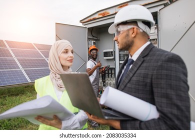 Male And Female Inspectors With Laptop And Papers Standing Talking On Field With Solar Panels. African American Technician Standing Behind Near Switchgear And Showing Thumb Up On Camera.