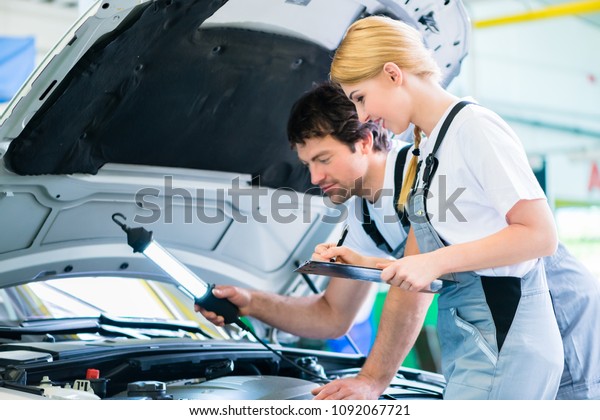 Male and female mechanic team\
examine car engine with light and checklist in workshop\
