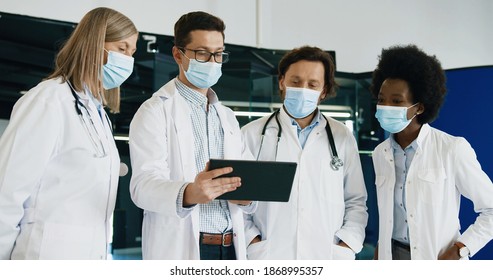 Male And Female Mixed-races Doctors In Medical Mask Discussing Coronavirus Disease While Typing On Tablet Standing In Hospital. Portrait Of Healthcare Professionals At Work. Covid 19 Pandemic