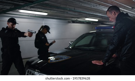 Male And Female Police Officers Arresting Robber. Dangerous Man Holding Hands On Patrol Car. Policeman Aiming Criminal With Gun At Detention. Multi Ethnic Police Team Working Together