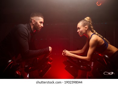 male and female sport people hold competition on bike in gym isolated on red neon lighted smoky space, oppsite of each other