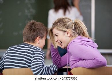 Male and a female student gossiping in classroom while the teacher solving a question on the blackboard.