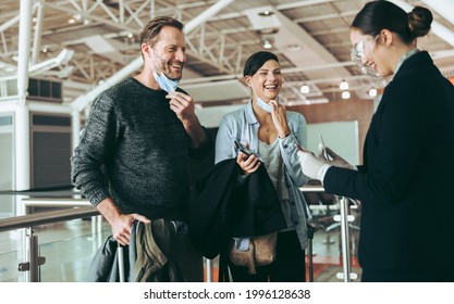 Male And Female Tourist With Face Masks Down At Departure Gate With Airport Staff In Face Shield. Flight Attendant Examining Passport And Boarding Pass Of Passengers At Airport Departure Gate.