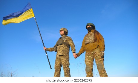 Male and female ukrainian army soldiers holding hands of each other and lifted up flag against blue sky. Military people waving flag of Ukraine in honor of the victory against Russian aggression. - Shutterstock ID 2256242595