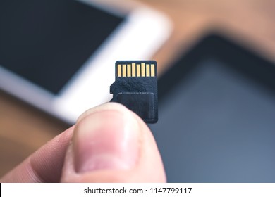 Male Fingers Holding A Micro SD Card With Tablet And Smartphone In Background, Extend Disk Space Concept