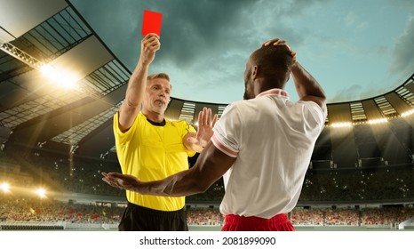 Male football referee showing soccer player a red card for breaking rules at crowded stadium over night cloudy sky. Concept of healthy lifestyle, sport, action, motion. Looks displeased.