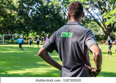 Male Football Or Soccer Coach Watching His Team Play At A Beautiful Soccer Field With Larger Background Trees