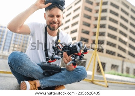 Male fpv pilot taking off goggles and checking his drone after landing on a street road. Operating multicopter experience, unmanned aviation concept. Focus is on the copter.
