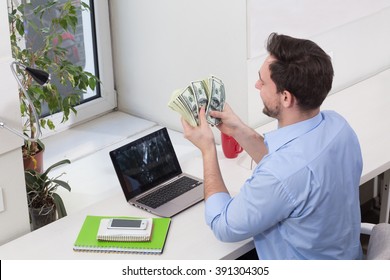 Male freelancer counting money or dollars in office while working on laptop or computer. Handsome man looking on money.