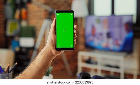Male freelancer holding smartphone with green screen display in living room, using isolated template with chroma key and mockup. Analyzing blank copyspace background on mobile phone. - Shutterstock ID 2230813895