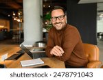 Male freelancer with laptop and notepad on desk smiling at camera while sitting on chair in office
