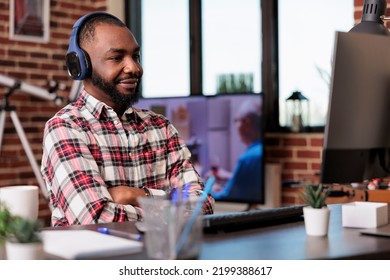 Male Freelancer Using Headphones And Working Remotely On Computer At Home Desk. Listening To Music Or Podcast Show Online On Social Media Internet, Attending Class Lesson Webinar.