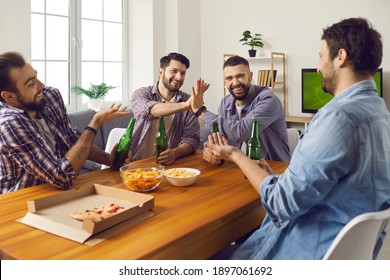 Male friends enjoying good time at fun get-together on weekend. Group of happy young men sitting at table at home, drinking beer, eating pizza, watching TV, playing games and telling funny stories