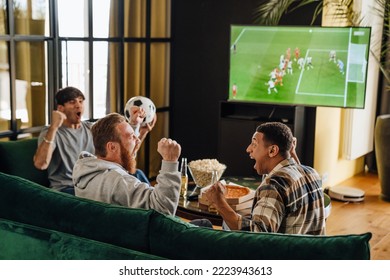 Male friends gesturing as winners while watching football match sitting on sofa in front of the TV screen - Shutterstock ID 2223943613