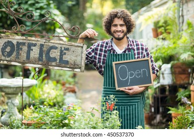 Male gardener holding open sign while standing by office placard outside greenhouse - Powered by Shutterstock