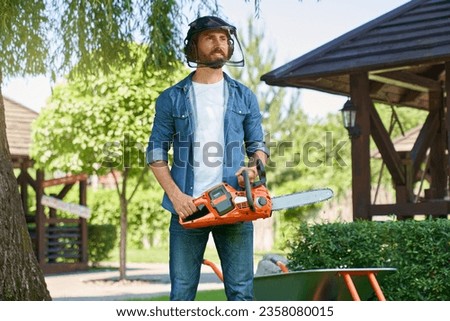 Male gardener in protective shield standing with cordless chain saw, working in summer garden. Front view of worker with beard holding modern electric saw, while looking ahead. Concept of gardening. 