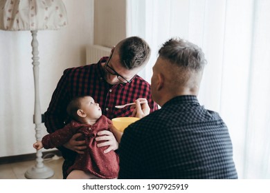 Male Gay Couple With Adopted Baby Girl At Home - Two Handsome Dads Feed The Baby Girl On Kitchen - Male Babysitters - Lgbt Family At Home - Diversity Concept