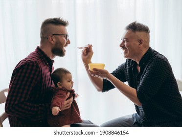 Male Gay Couple With Adopted Baby Girl At Home - Two Handsome Dads Feed The Baby Girl On Kitchen - Male Babysitters - Lgbt Family At Home - Diversity Concept