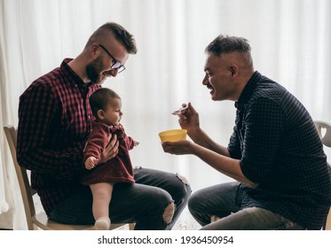 Male gay couple with adopted baby girl at home - Two handsome dads feed the baby girl on kitchen - Male babysitters - Lgbt family at home - Diversity concept
