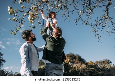 Male Gay Couple Lifting Their Young Daughter To Touch The Flowers Of A Tree. Modern Family Concept.