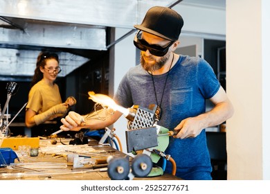 A male glassblower wearing safety glasses and a cap works on shaping a wine glass with a blowtorch in a studio setting. A female one watches in the background. - Powered by Shutterstock