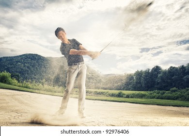 Male golf player in blue shirt and grey pants hitting golf ball out of a sand trap with sand wedge and sand caught in motion. - Powered by Shutterstock