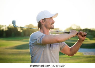 Male golf player isolated on beautiful sunset. Smiling golfer with white hat on holding golf club over shoulder.