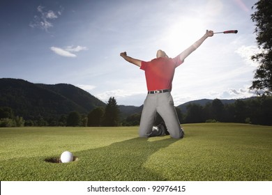 Male Golf Player On Knees And Arms Raised With Putter In Hand In Winner Pose On Golf Green Being Overjoyed As Golf Ball Drops Into Cup.